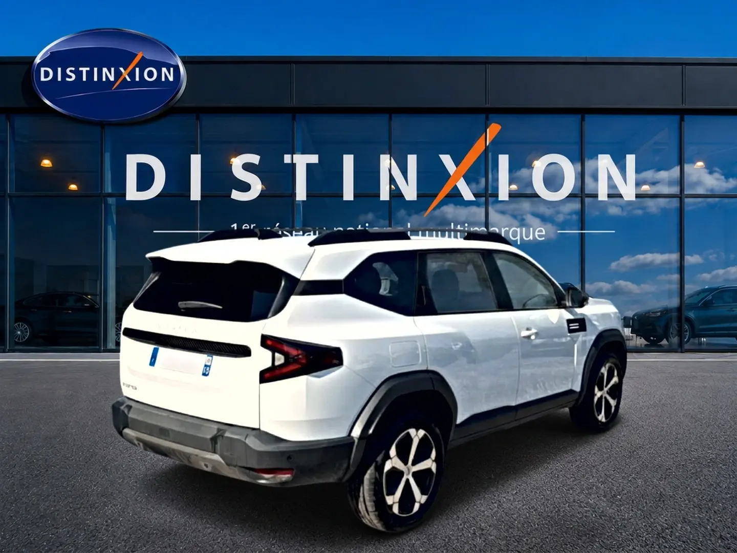 Vue 3/4 arrière droite du SUV blanc Dacia Bigster 2026 avec jantes alu et toit noir devant une vitrine Distinxion.