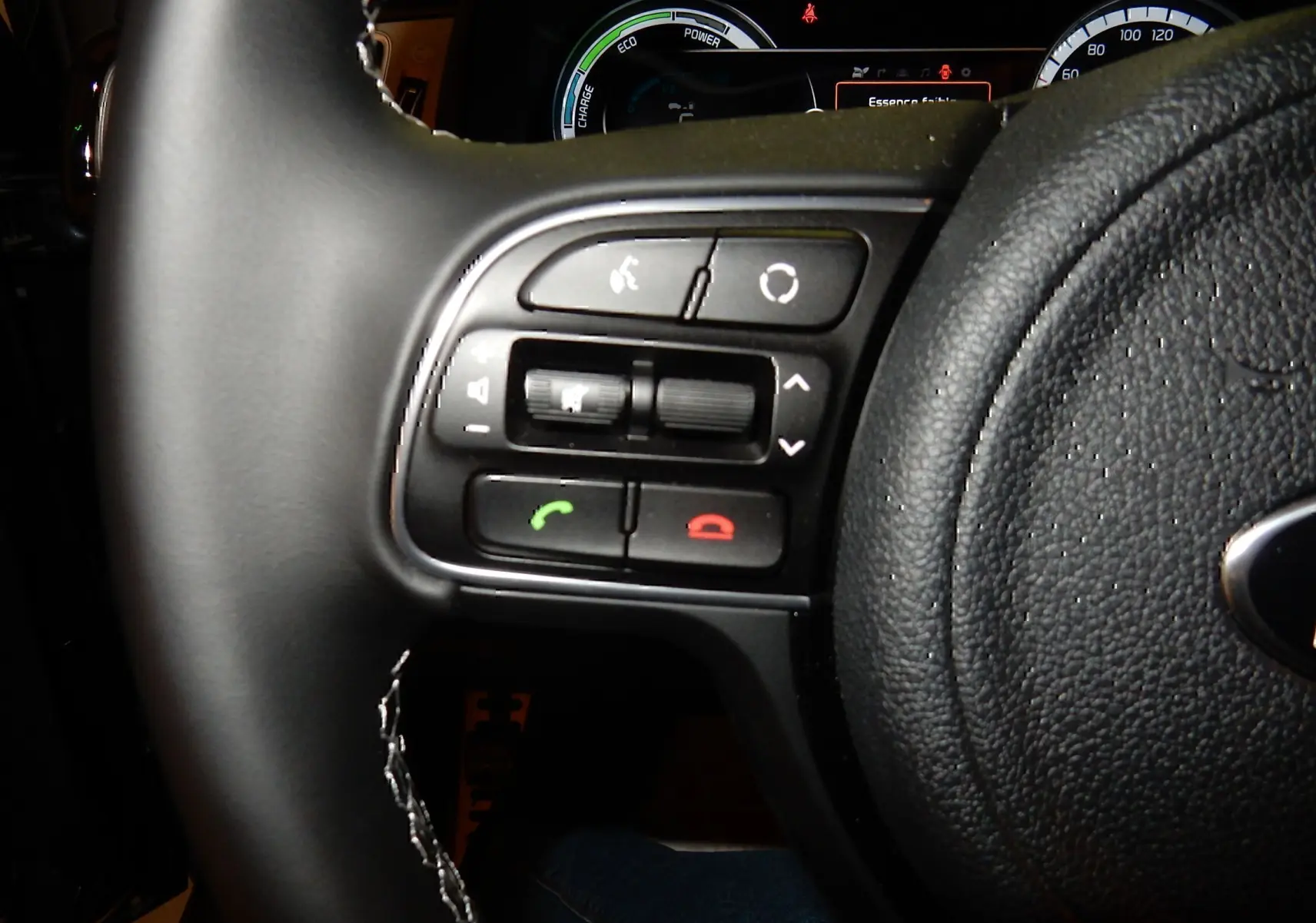 Gros plan sur les commandes gauche du volant noir du Kia Niro hybride gris galène, avec tableau de bord numérique en arrière-plan.