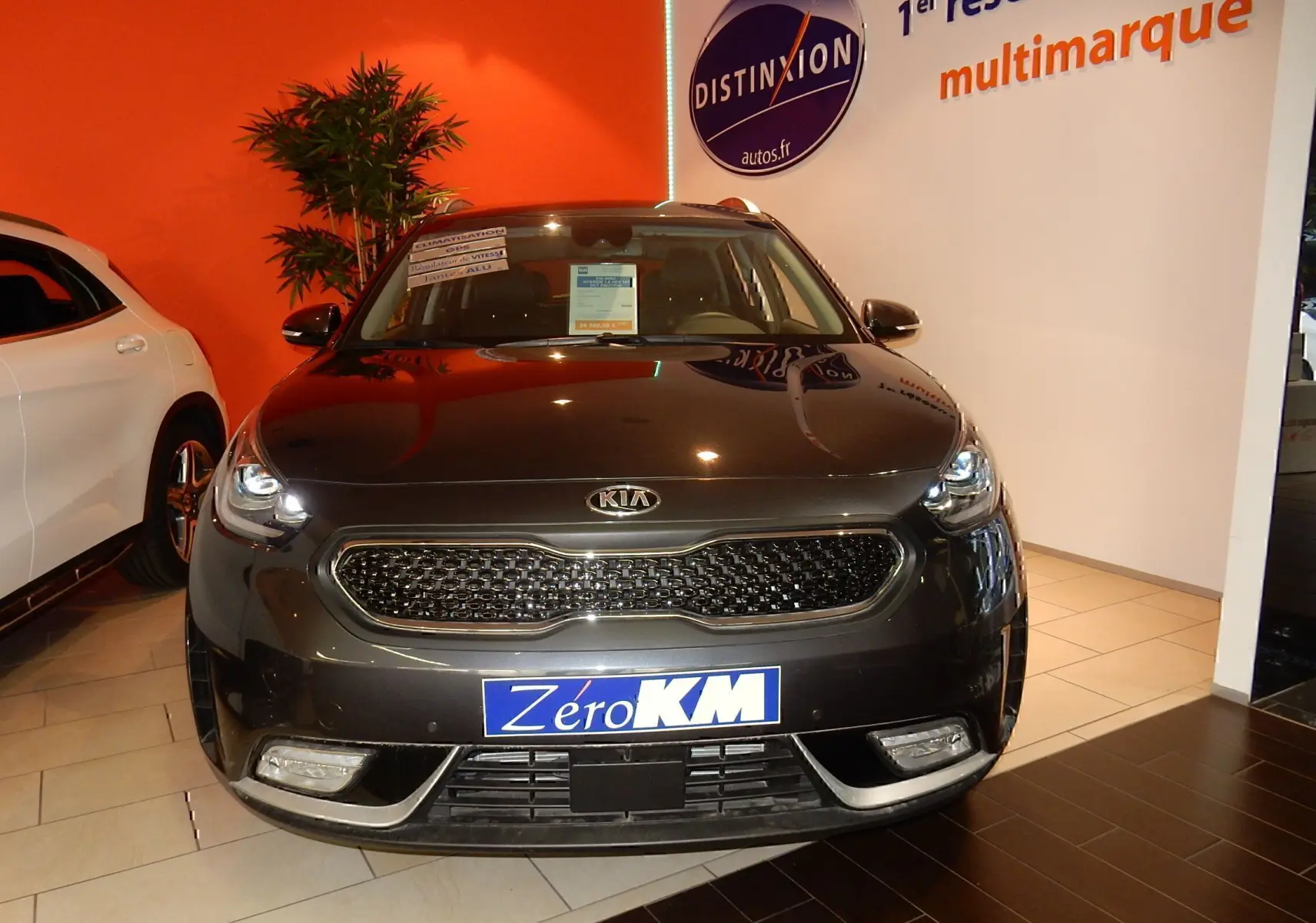 Vue avant d'un KIA Niro hybride gris galène métal exposé en showroom avec plaque Zéro KM visible.