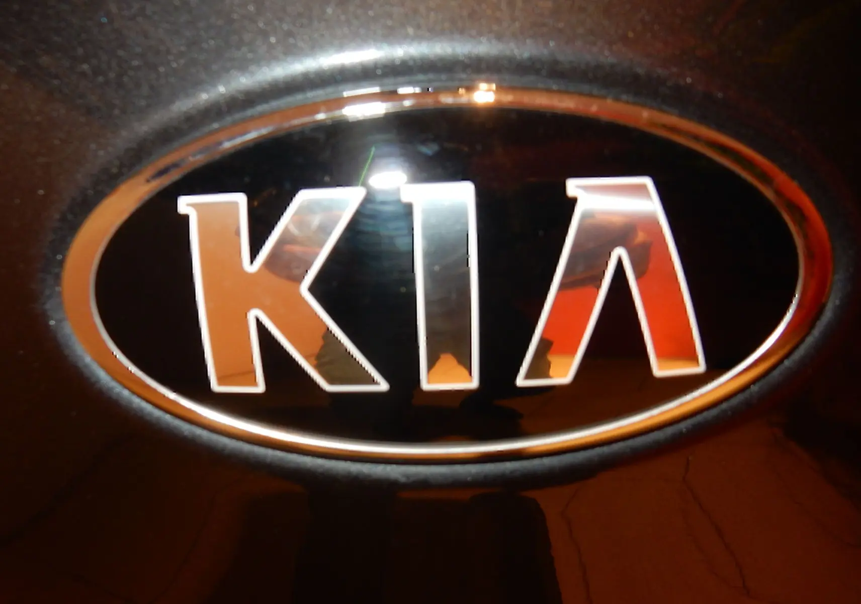 Gros plan sur le logo KIA en chrome sur fond gris galène métallique, éclairé par une lumière chaude.