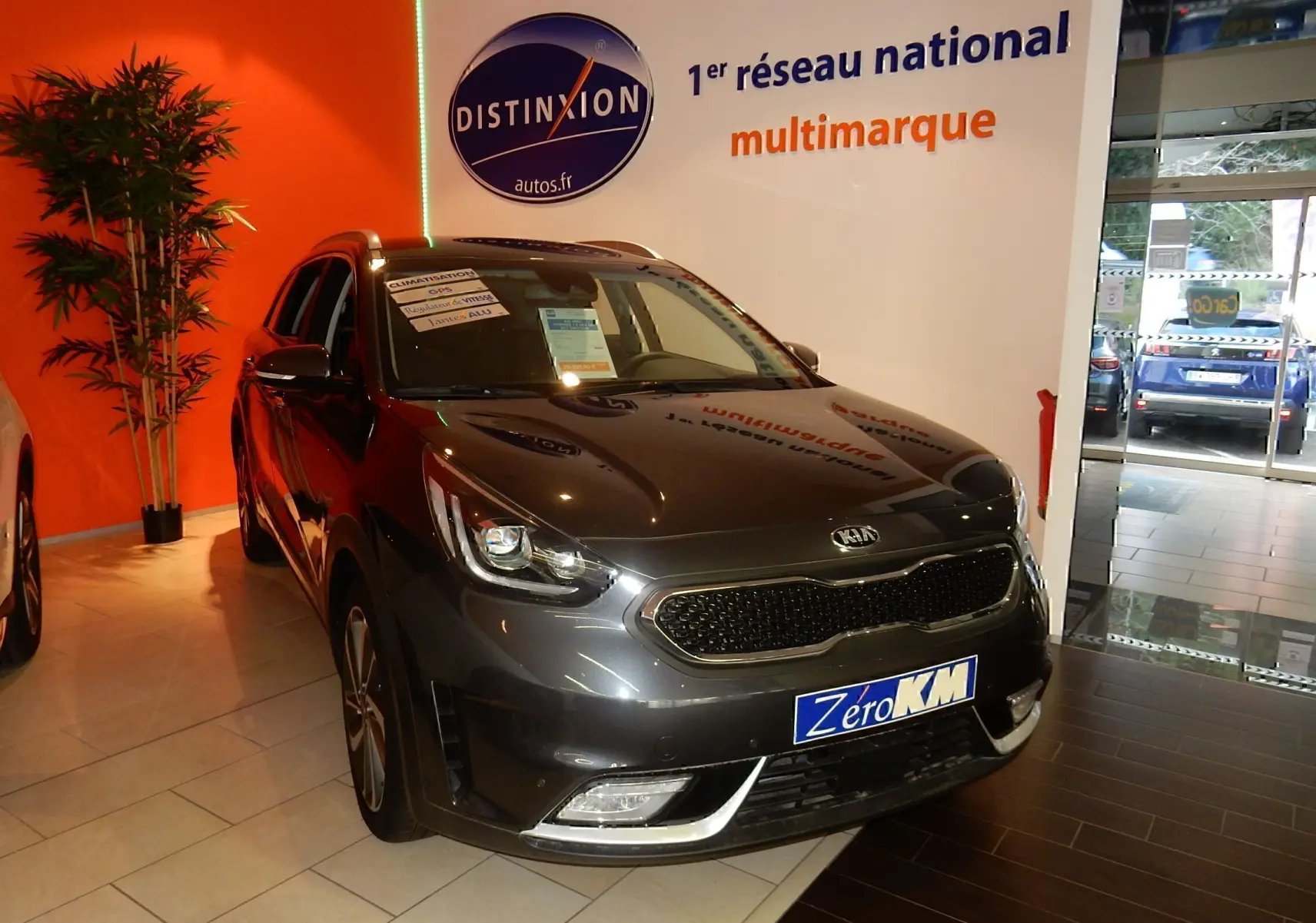 KIA Niro hybride gris galène en 3/4 avant droit, stationné en intérieur avec panneau Distinxion en arrière-plan.