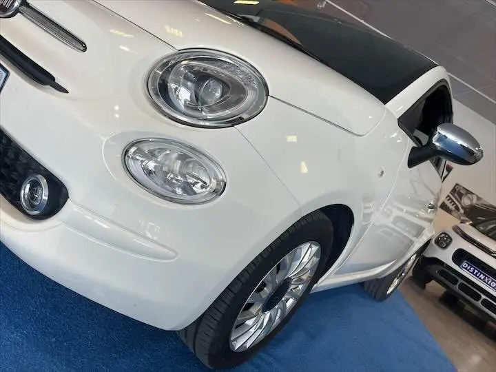 Vue 3/4 avant gauche d'une FIAT 500 blanche avec toit noir et jantes argentées en showroom.