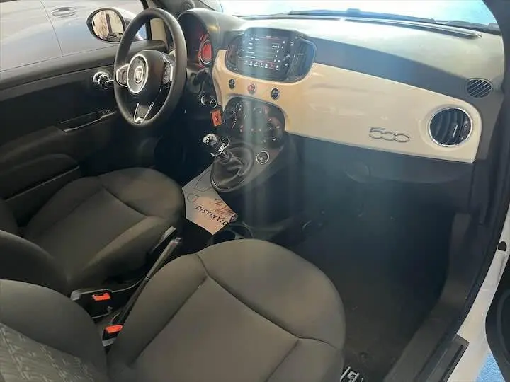 Vue intérieure côté conducteur de la Fiat 500 blanche 2023, tableau de bord beige avec logo 500 et boîte manuelle.