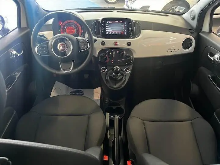 Vue intérieure avant de la Fiat 500 blanche 2023, tableau de bord brillant avec écran tactile et volant multifonction noir.