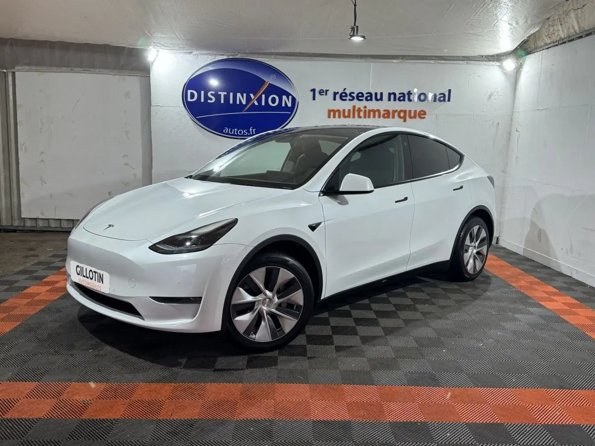 Tesla Model Y blanc en 3/4 avant droit dans un showroom avec sol noir et orange et logo Distinxion en arrière-plan