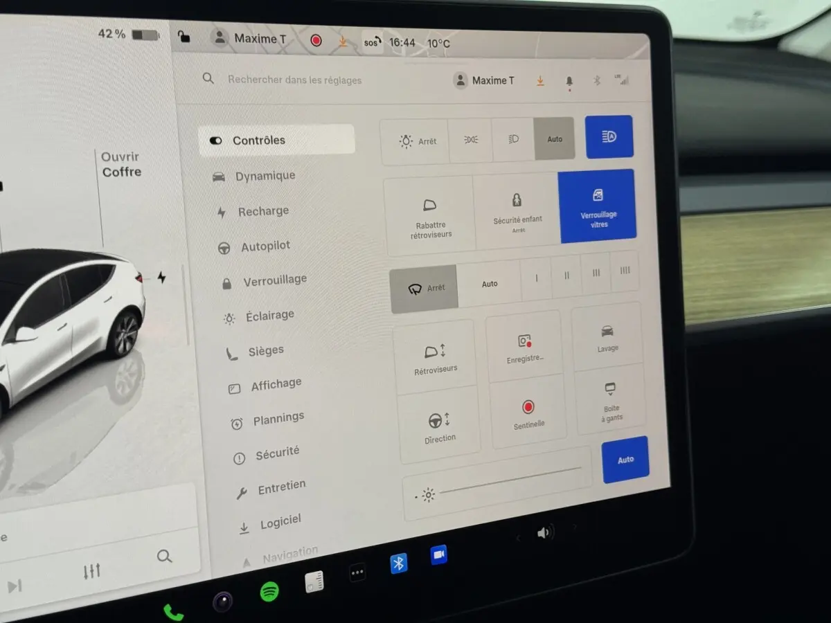 Écran tactile central affichant les réglages du Tesla Model Y blanc, intérieur avec tableau de bord bois clair.