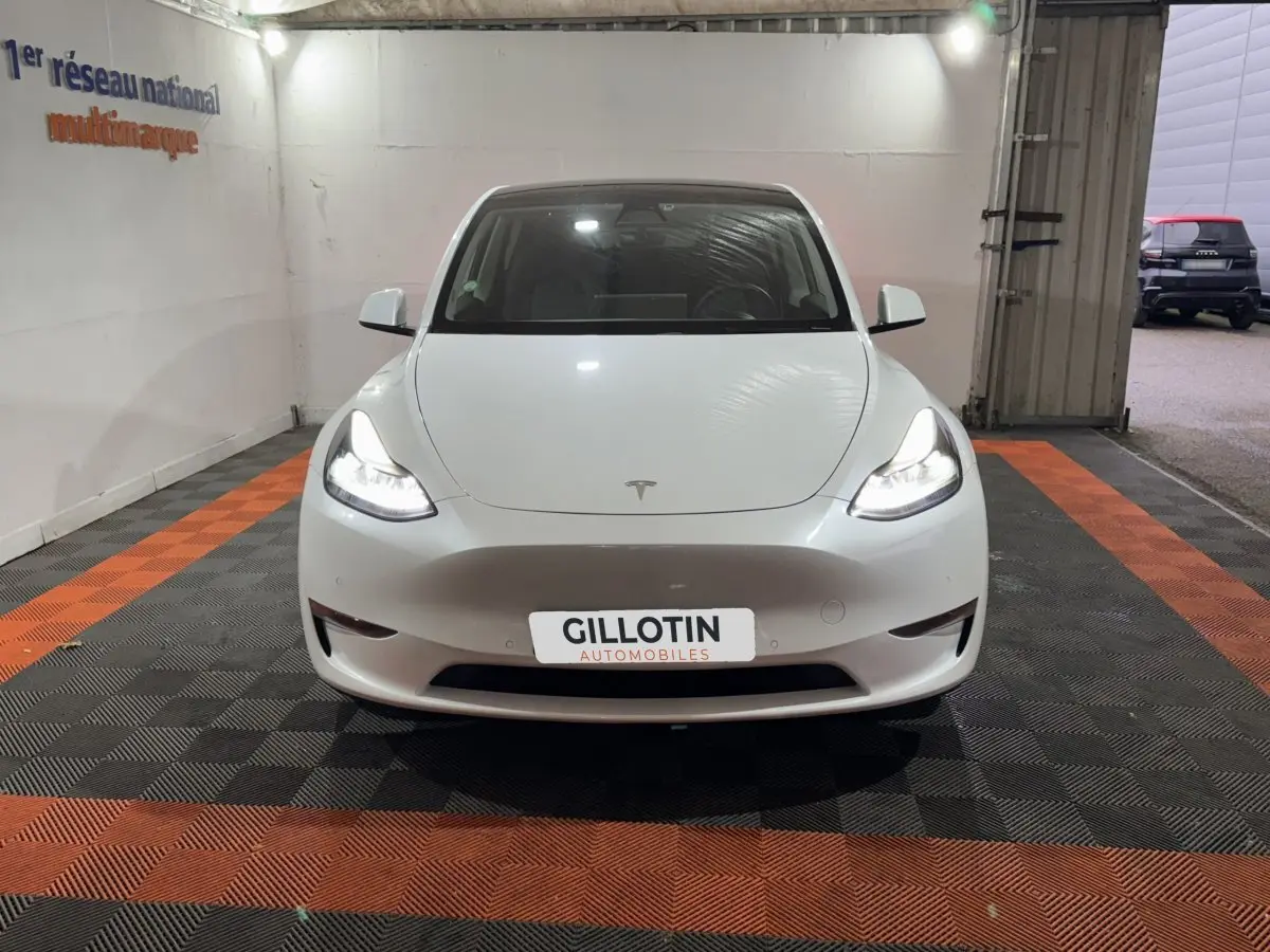 Tesla Model Y blanc en vue frontale dans un garage avec phares allumés et plaque GILLOTIN Automobiles.