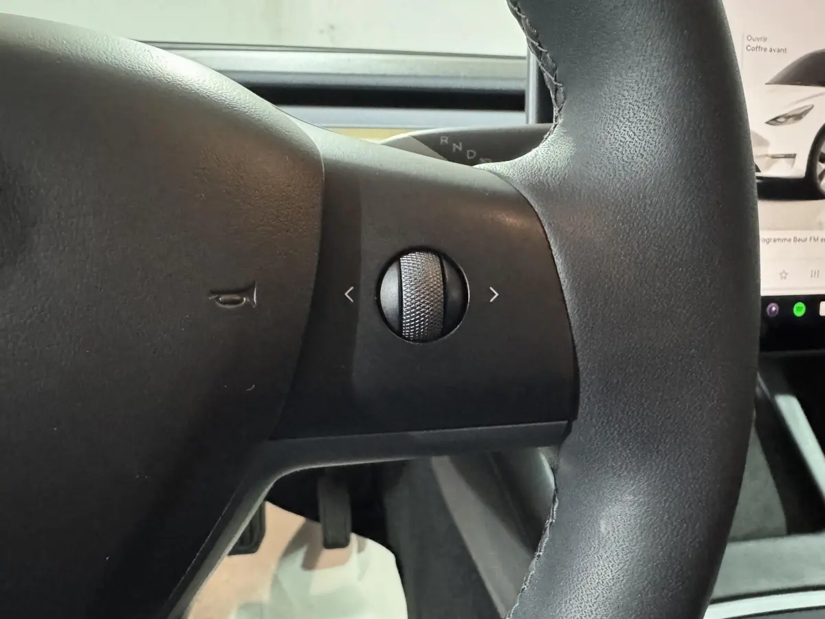 Gros plan sur la molette de commande à droite du volant cuir noir d'une Tesla Model Y blanche, écran tactile visible en arrière-plan.