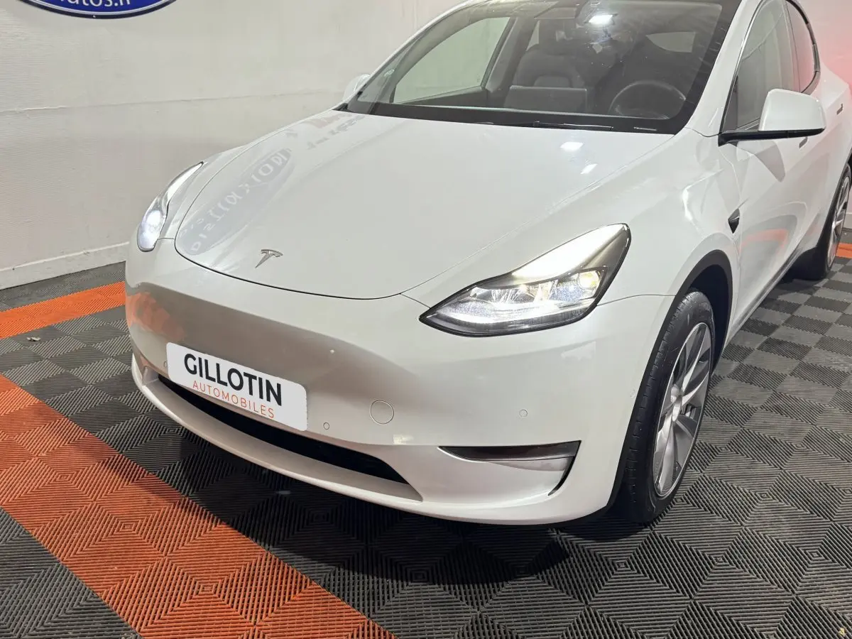 Vue 3/4 avant droit d'une Tesla Model Y blanche avec phares LED allumés en intérieur showroom.
