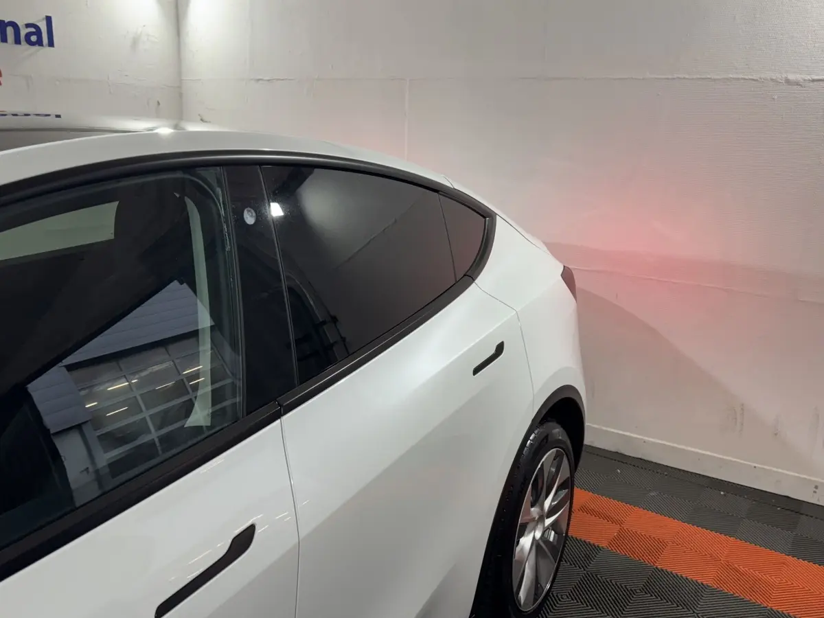 Vue 3/4 arrière côté gauche d'une Tesla Model Y blanche avec vitres teintées et jantes argentées dans un garage.