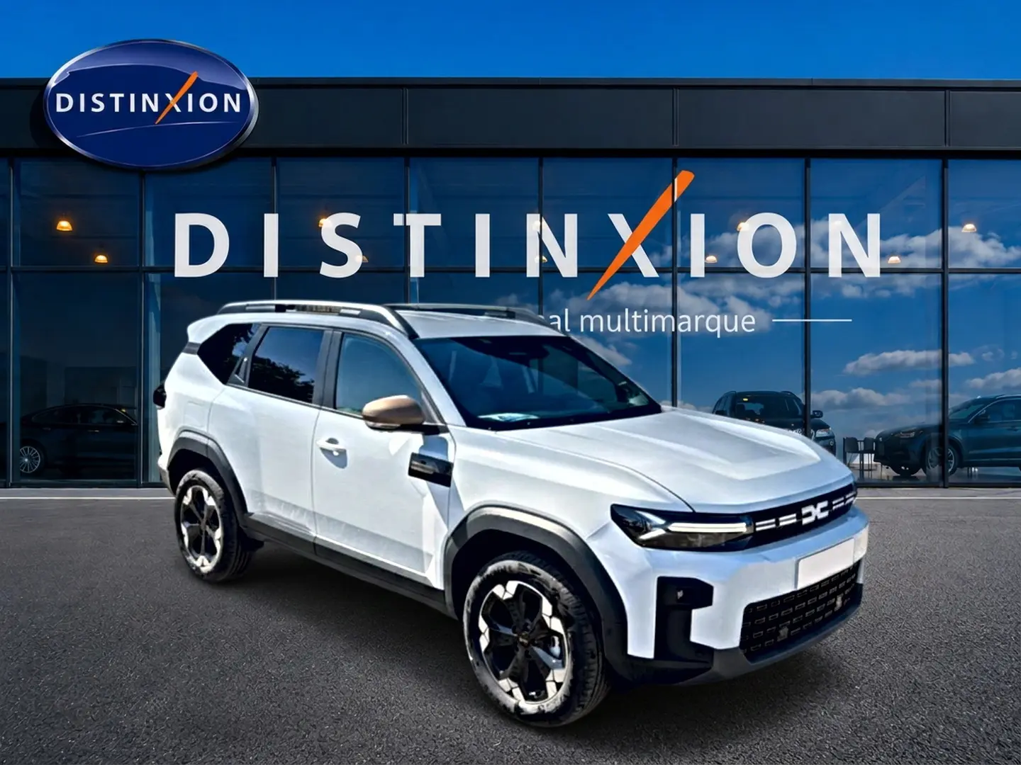 SUV Dacia Bigster blanc en 3/4 avant droit avec jantes noires et barres de toit, devant une vitrine Distinxion.