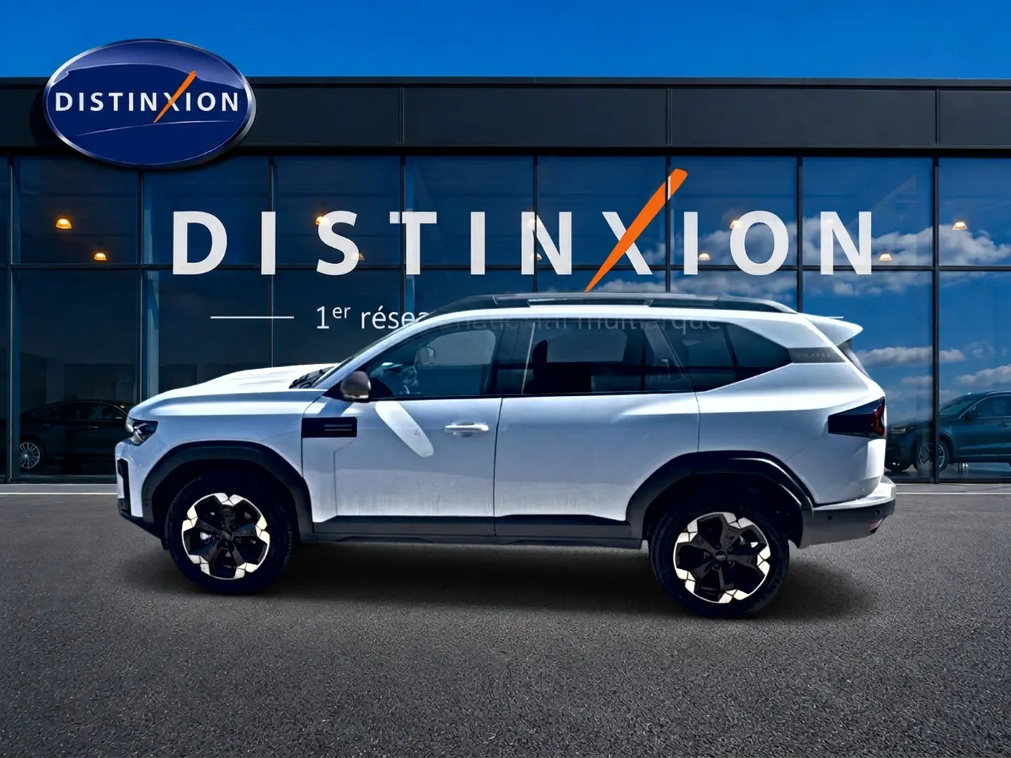 Profil latéral gauche du Dacia Bigster blanc 2026 avec jantes alu et barres de toit devant une vitrine Distinxion.