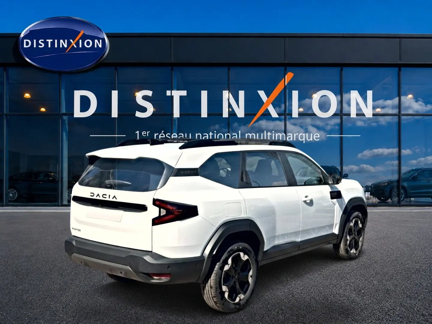 Vue 3/4 arrière droite du Dacia Bigster blanc avec barres de toit et jantes alu noires devant une vitrine Distinxion.
