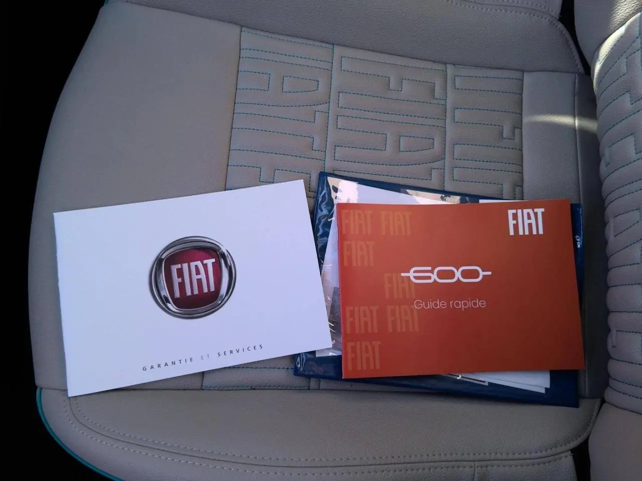 Vue rapprochée des manuels FIAT posés sur le siège avant beige avec surpiqûres bleues du Fiat 600 hybride blanc 2025.