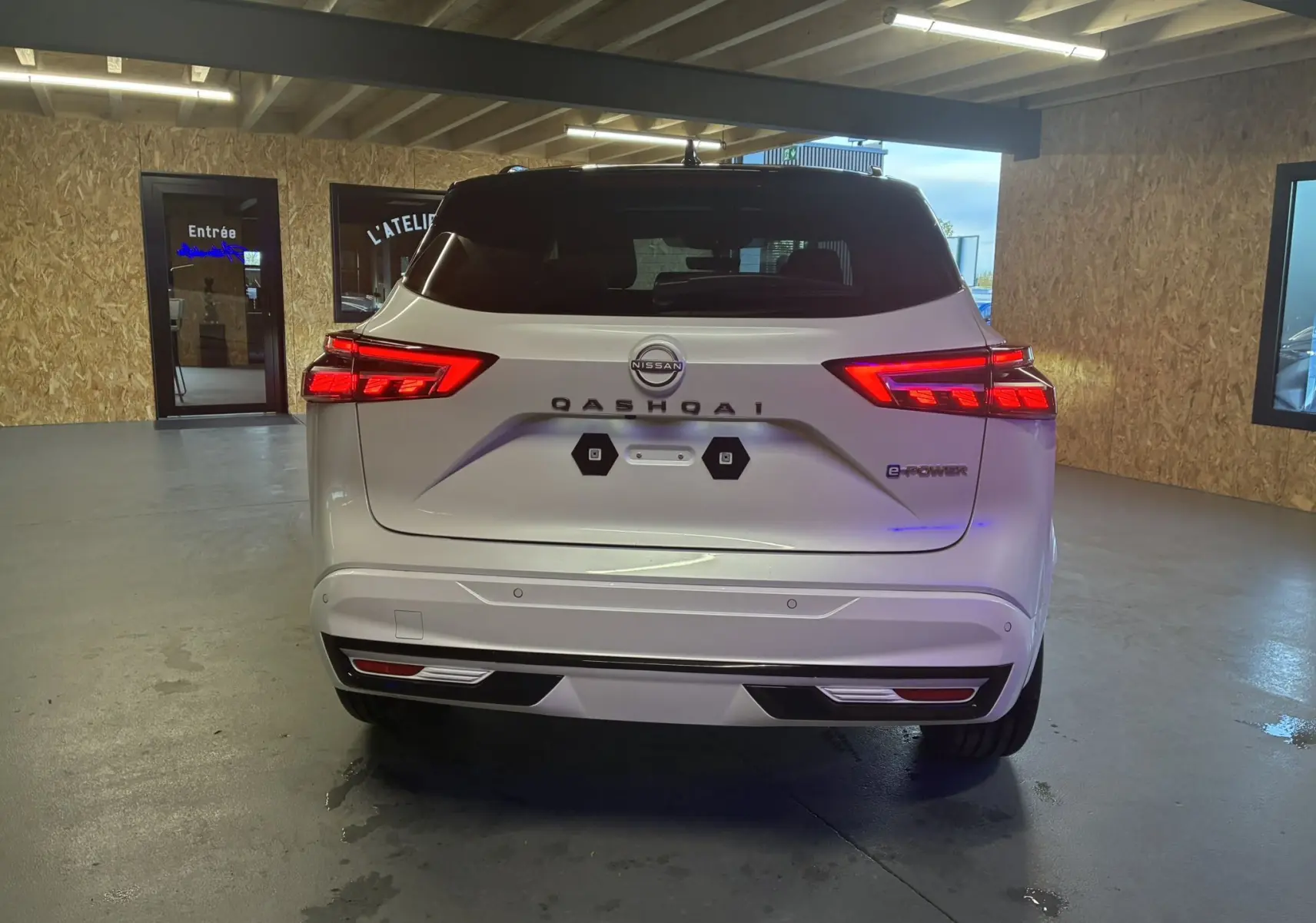 Vue arrière d'un Nissan Qashqai blanc nacré avec toit noir, feux LED allumés dans un garage intérieur.