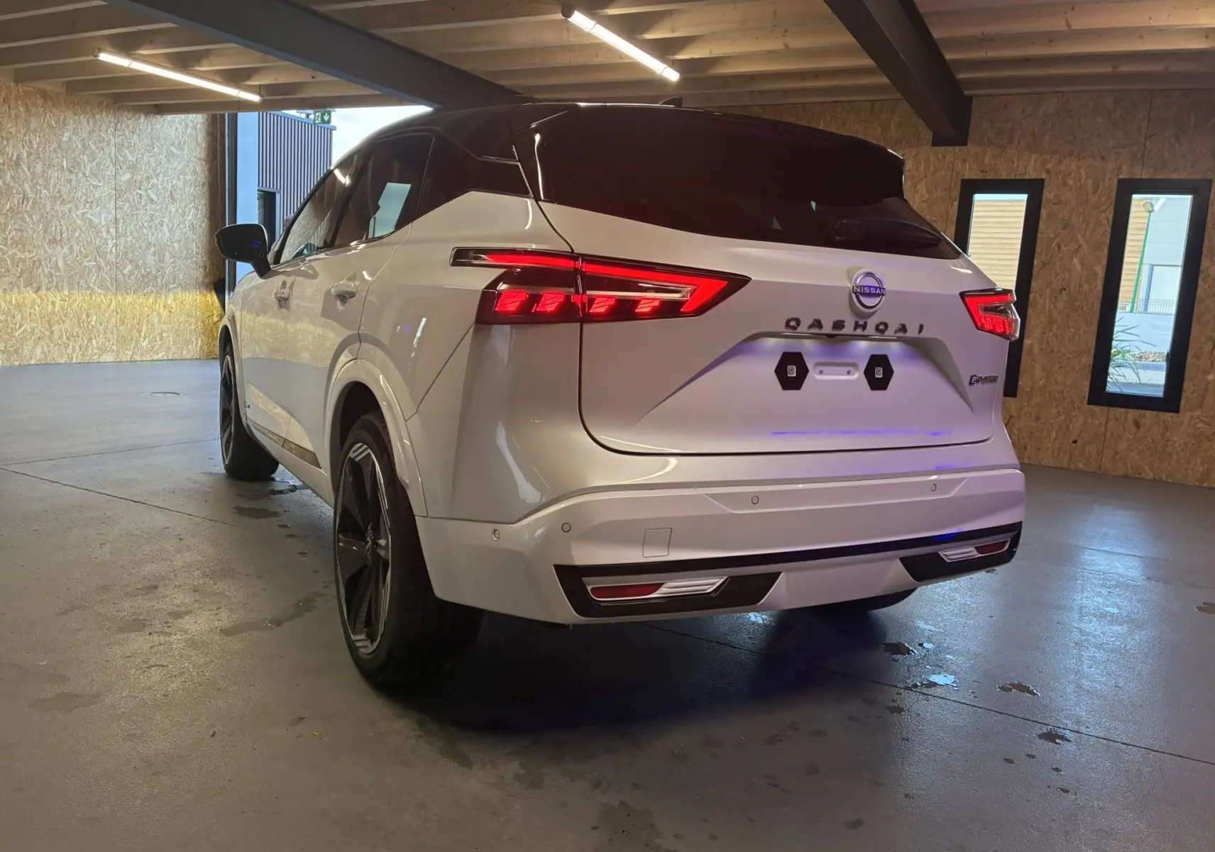 Vue 3/4 arrière d'un Nissan Qashqai 2025 blanc nacré avec toit noir, feux arrière LED et jantes noires dans un garage.