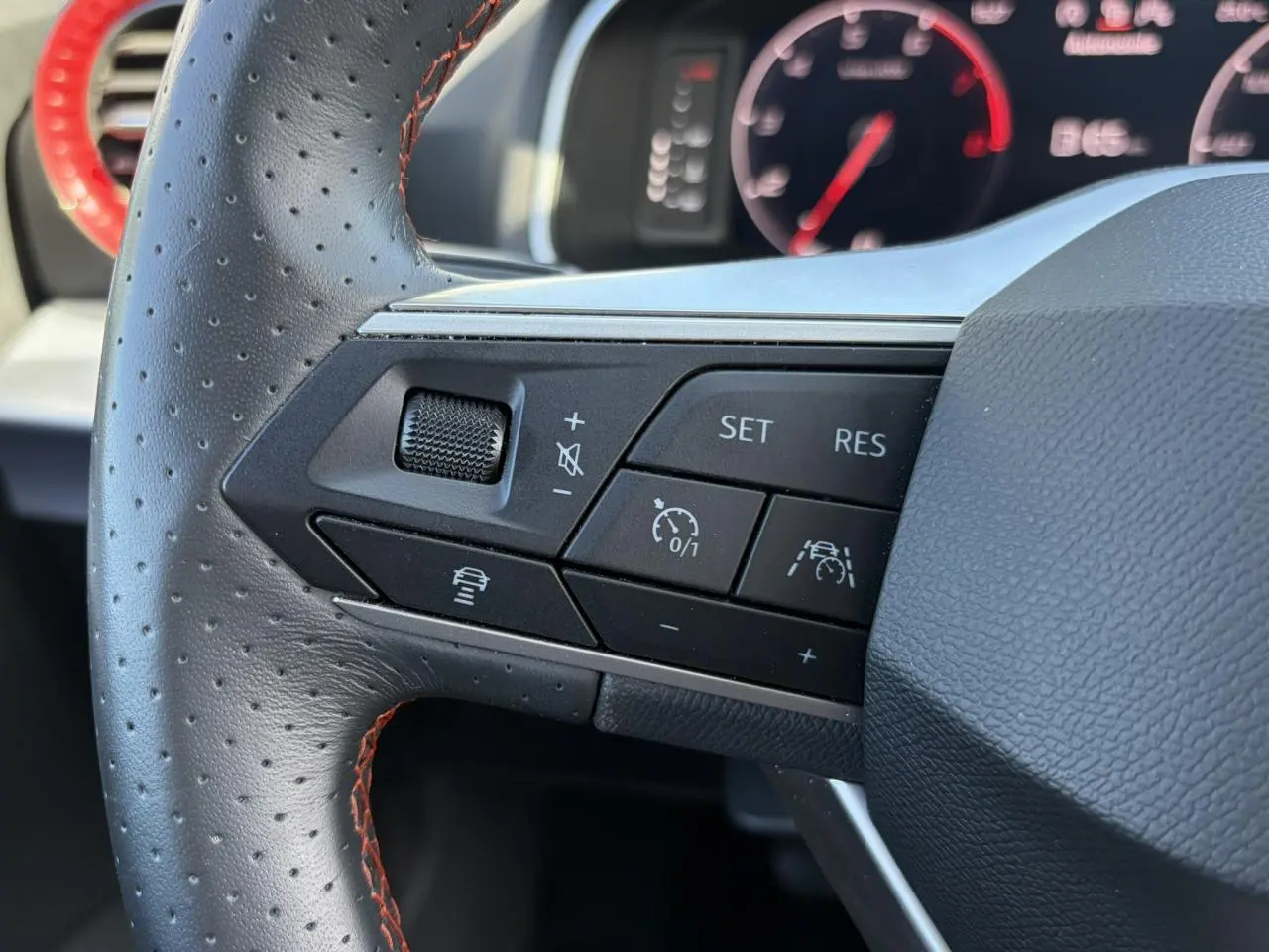 Gros plan sur les commandes à gauche du volant en cuir perforé noir avec surpiqûres rouges du SEAT Ibiza blanc Candy 2025.