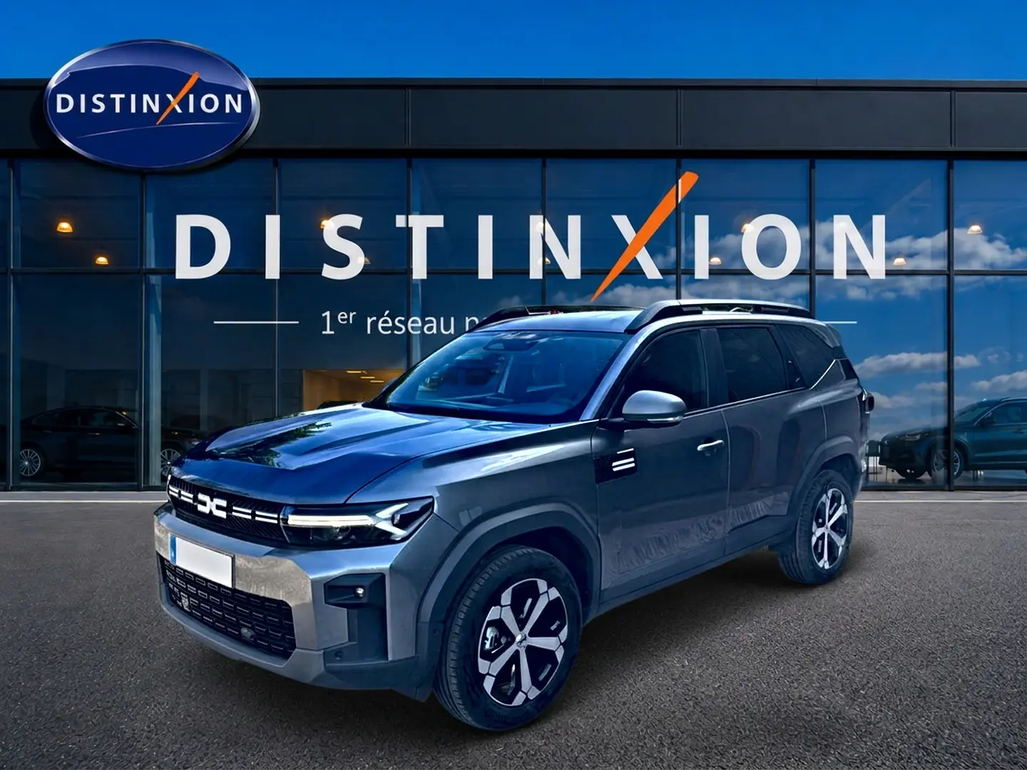Dacia Bigster gris schiste en 3/4 avant droit, avec jantes alu et barres de toit devant un showroom Distinxion.
