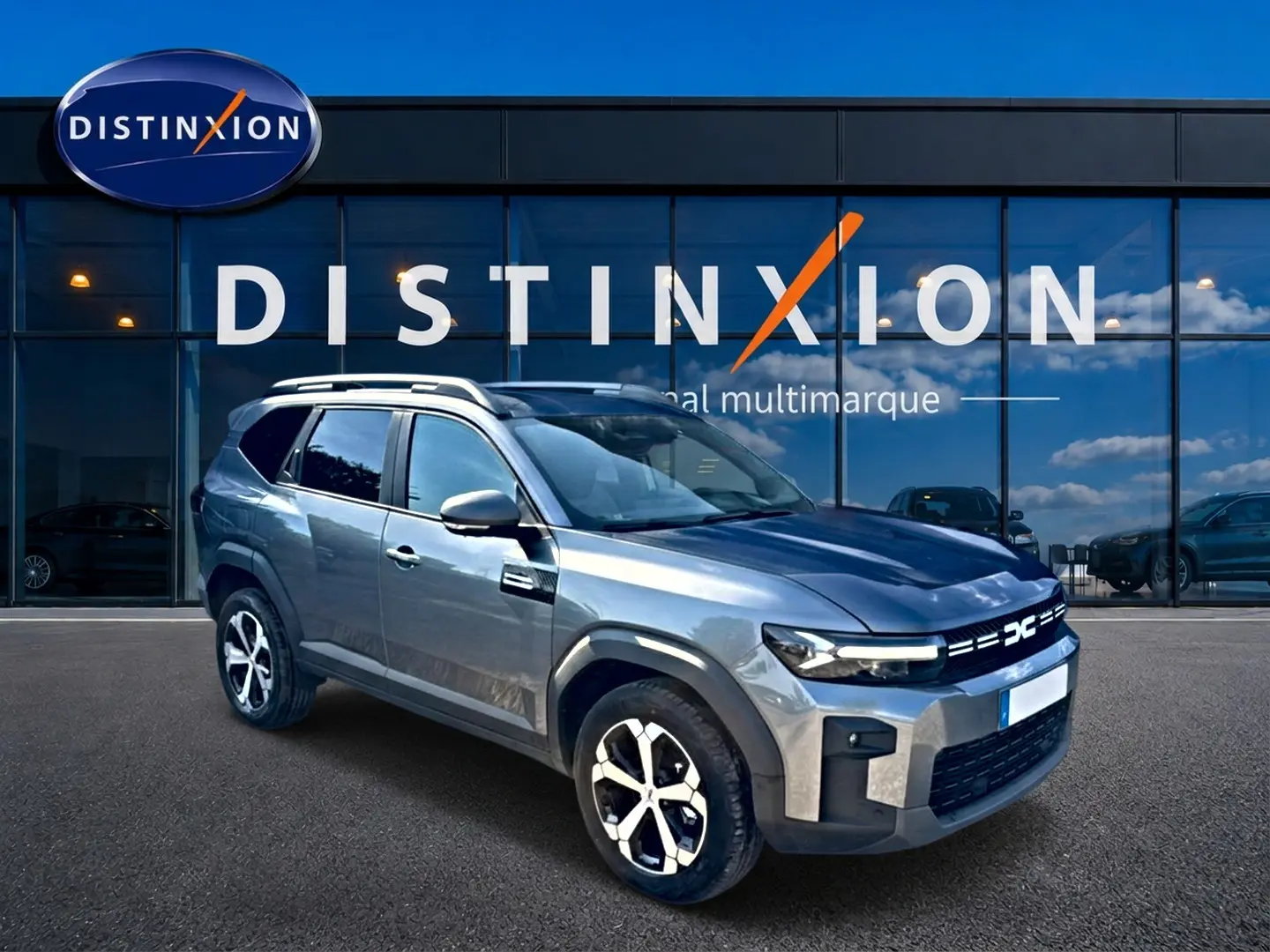 Dacia Bigster gris schiste en 3/4 avant droit, avec barres de toit et jantes alu distinctives, devant un showroom.