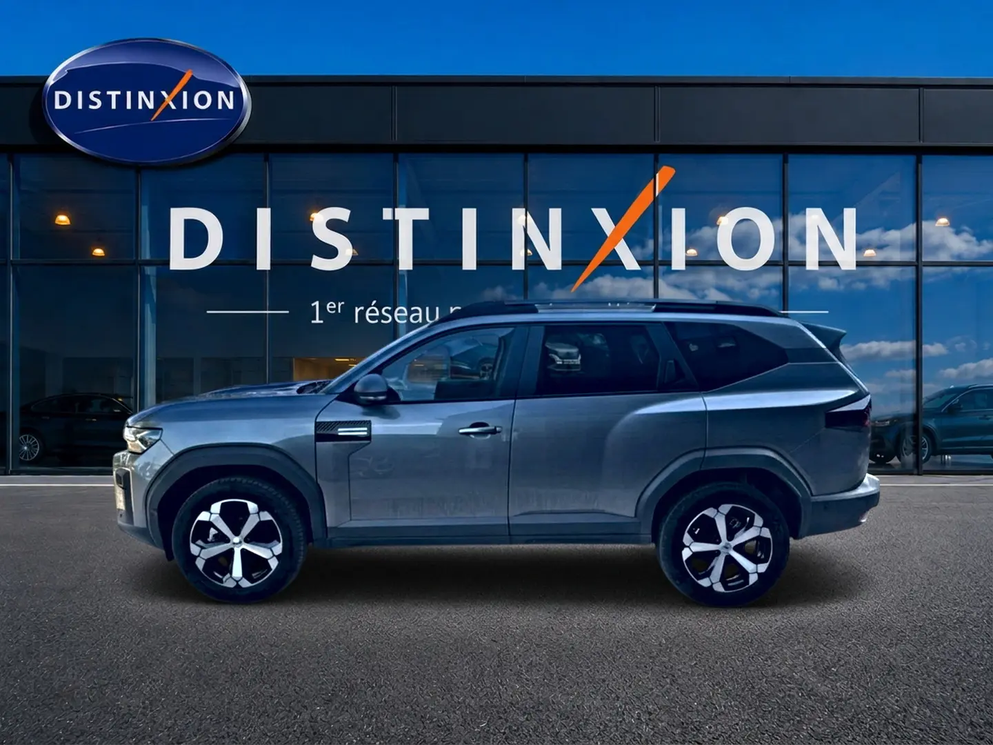 Profil côté gauche du Dacia Bigster gris schiste 2026 avec jantes alu et barres de toit visibles devant une vitrine Distinxion.