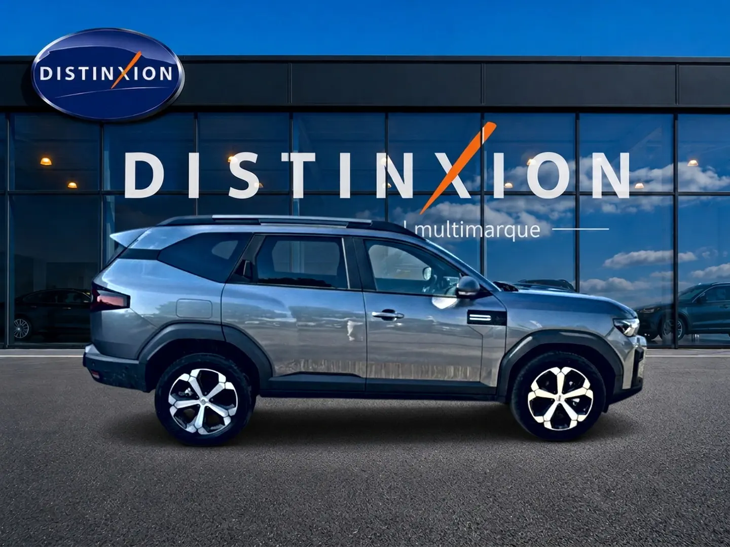 Profil droit du Dacia Bigster gris schiste 2026 avec jantes alu et barres de toit, devant une vitrine Distinxion.