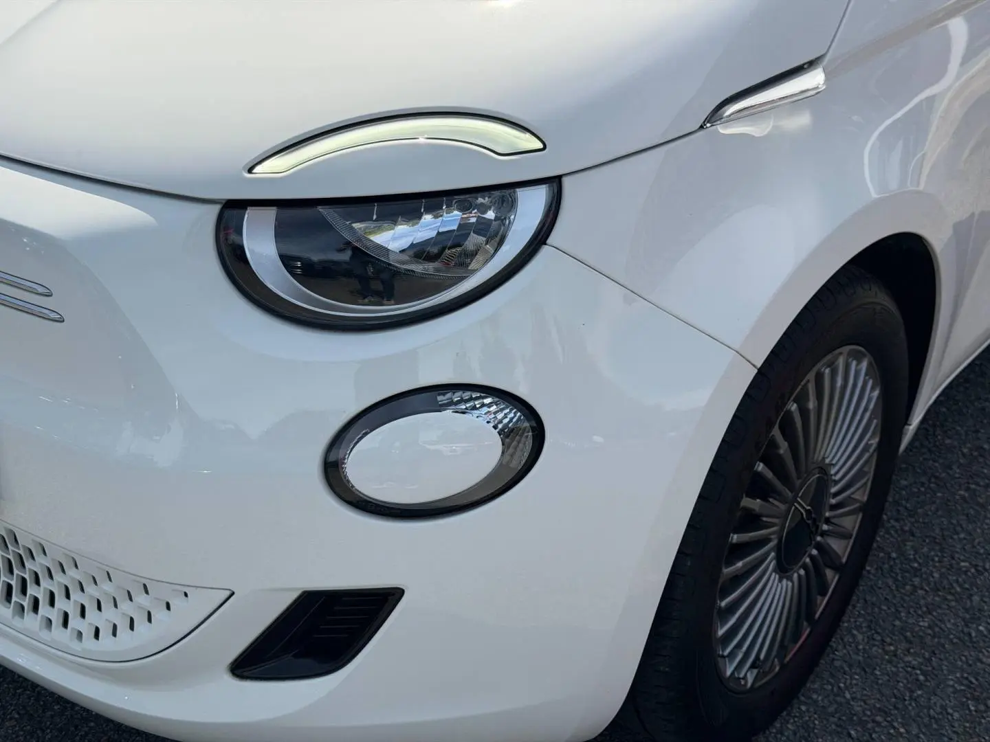 Gros plan sur l'avant droit blanc nacré du Fiat 500 e 118ch MY23, mettant en valeur le phare LED et la jante spécifique.