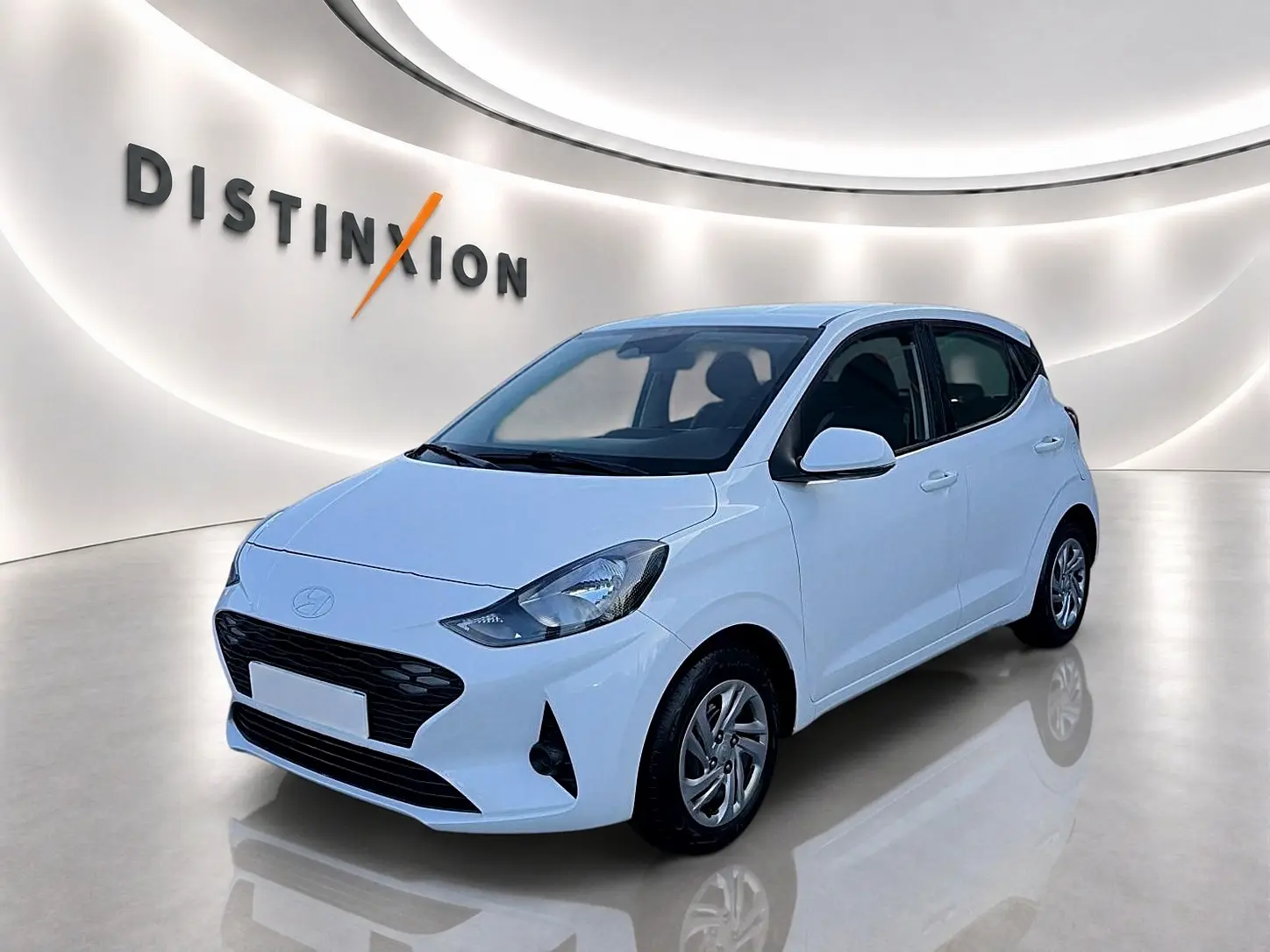 Vue 3/4 avant d'une Hyundai i10 blanche 2024 avec enjoliveurs et rétroviseurs assortis en intérieur showroom lumineux.