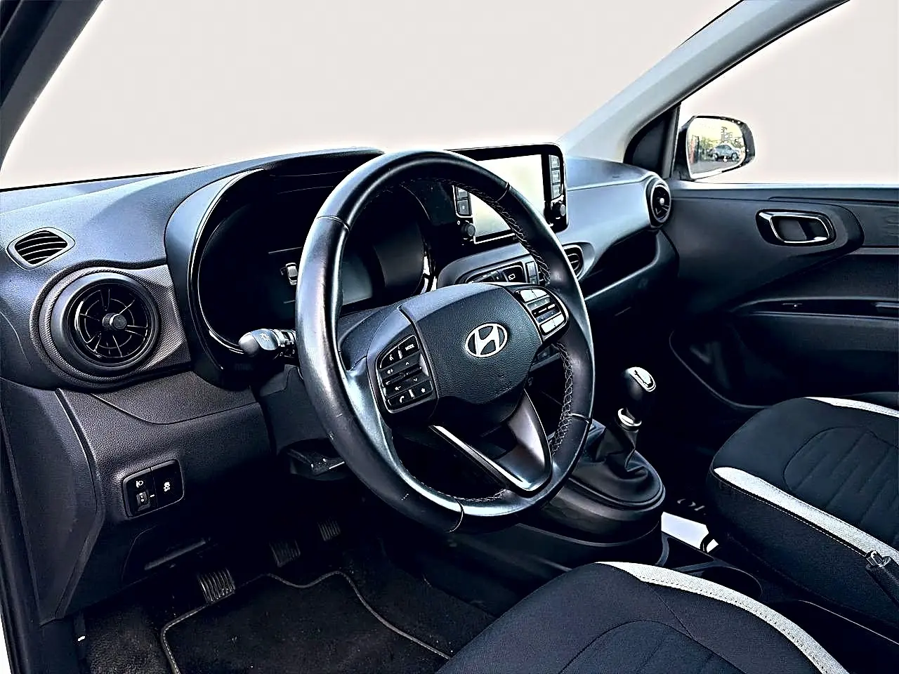 Vue intérieure côté conducteur du tableau de bord et volant cuir noir de la Hyundai i10 2024, avec levier de vitesses manuel.