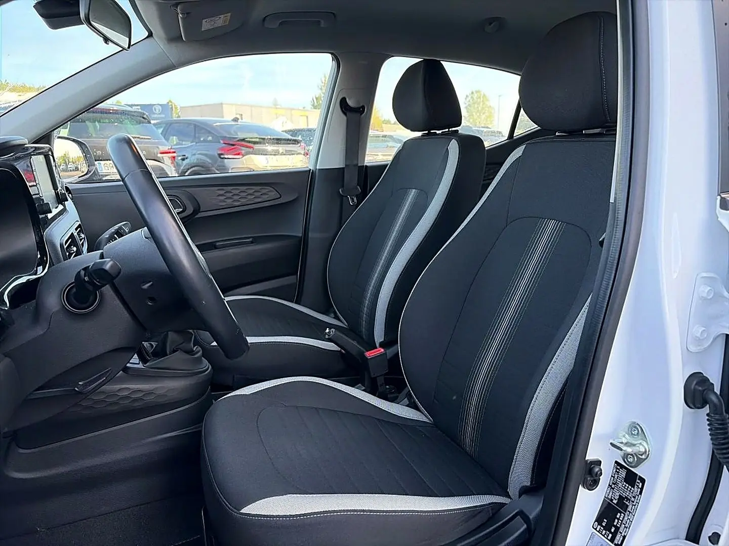 Vue intérieure côté gauche de la Hyundai i10 blanche 2024, montrant les sièges noirs avec surpiqûres grises et volant cuir multifonction.