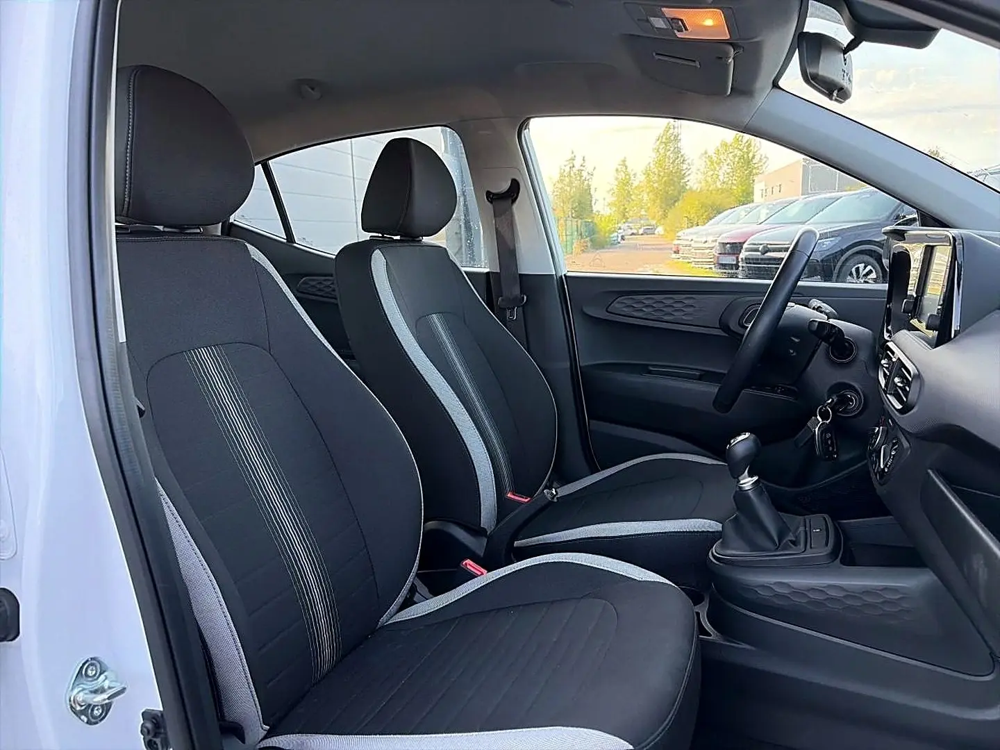 Vue intérieure côté gauche de la Hyundai i10 2024 blanche, montrant les sièges noirs avec surpiqûres grises et la console centrale.