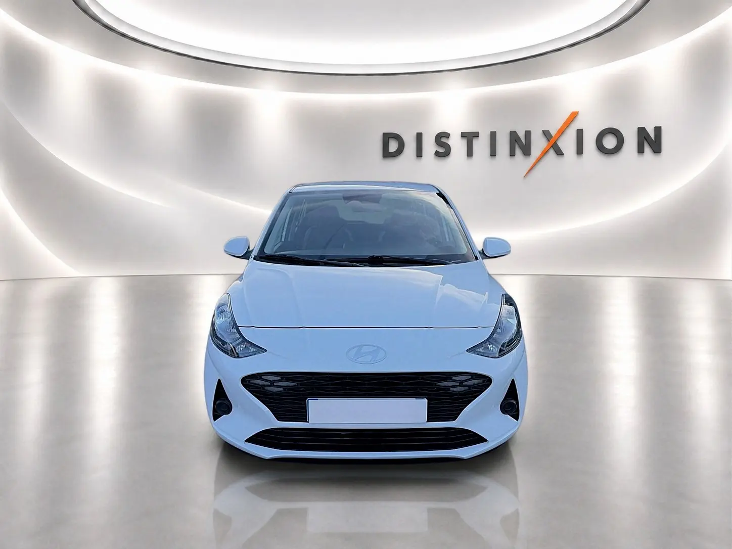 Vue avant d'une Hyundai i10 blanche 2024 dans un showroom lumineux avec logo Distinxion en arrière-plan.