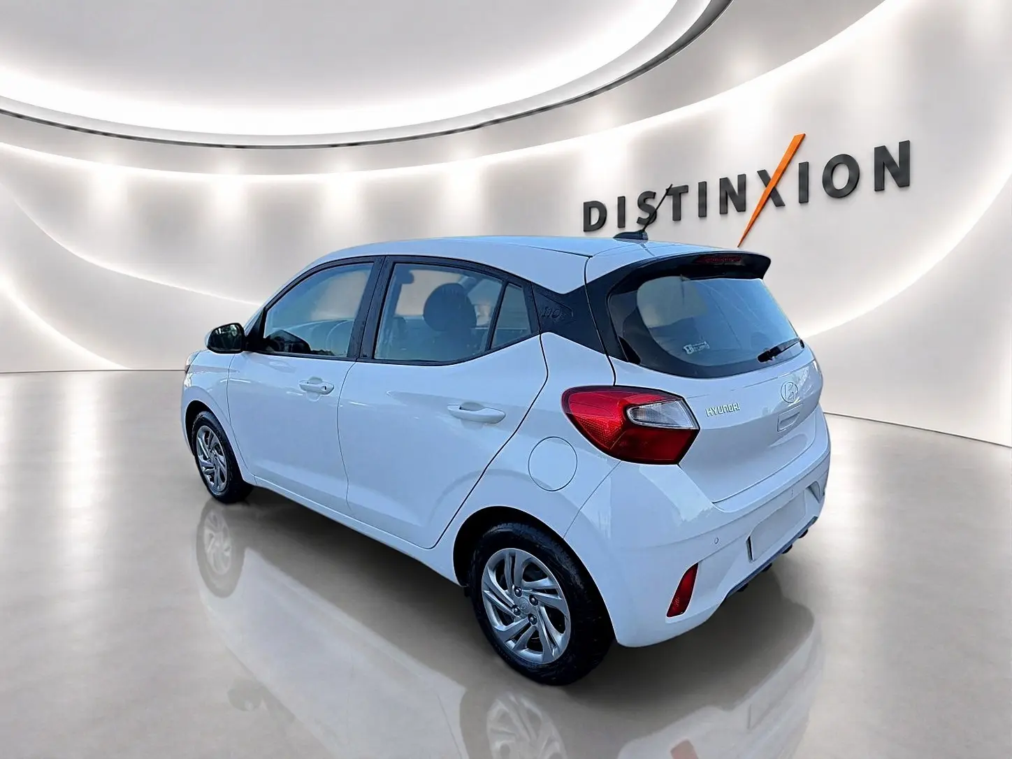 Vue 3/4 arrière droite d'une Hyundai i10 blanche dans un showroom lumineux avec logo Distinxion en arrière-plan.