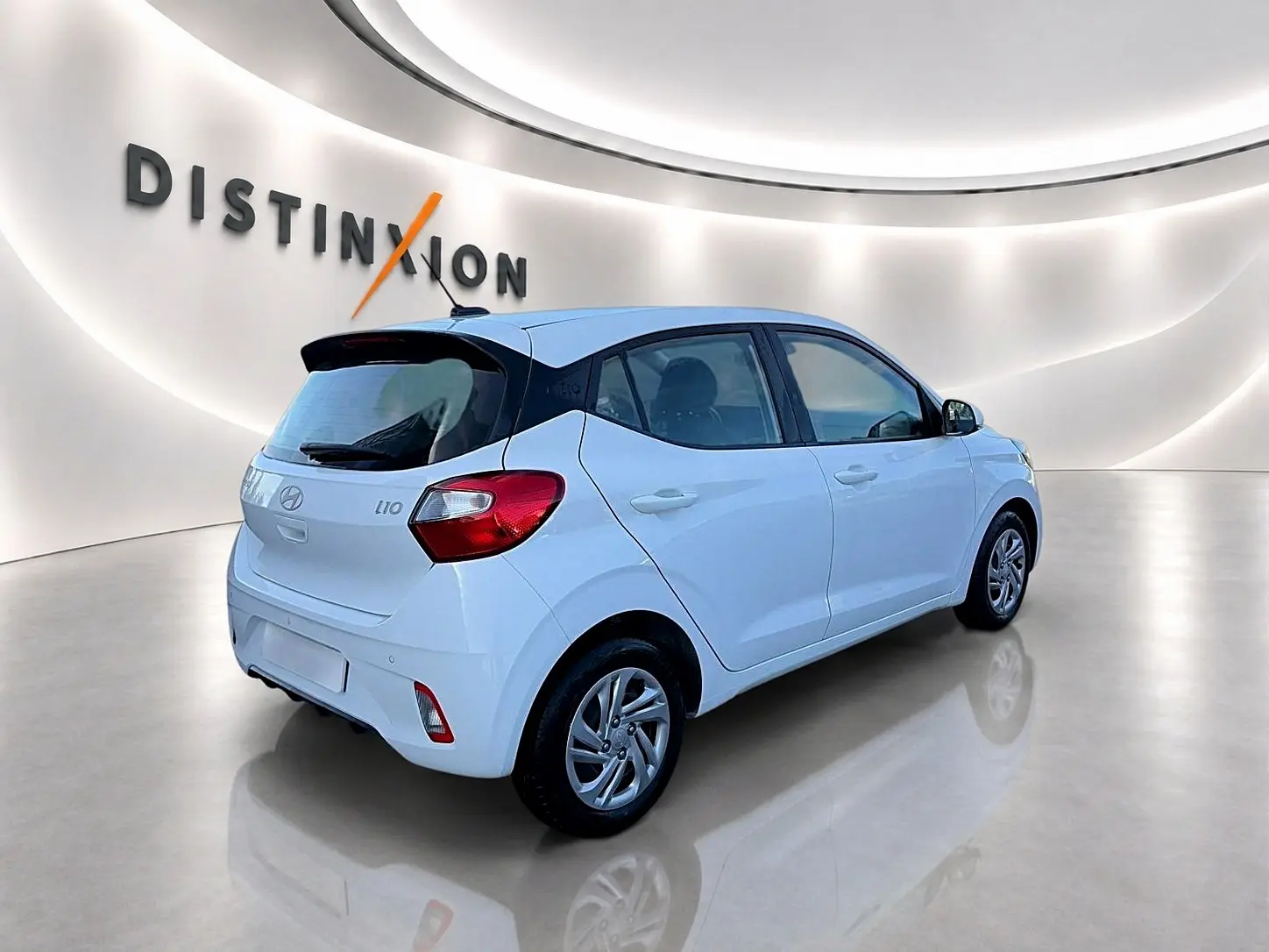 Vue 3/4 arrière droite d'une Hyundai i10 blanche 2024 dans un showroom lumineux avec logo Distinxion en arrière-plan.