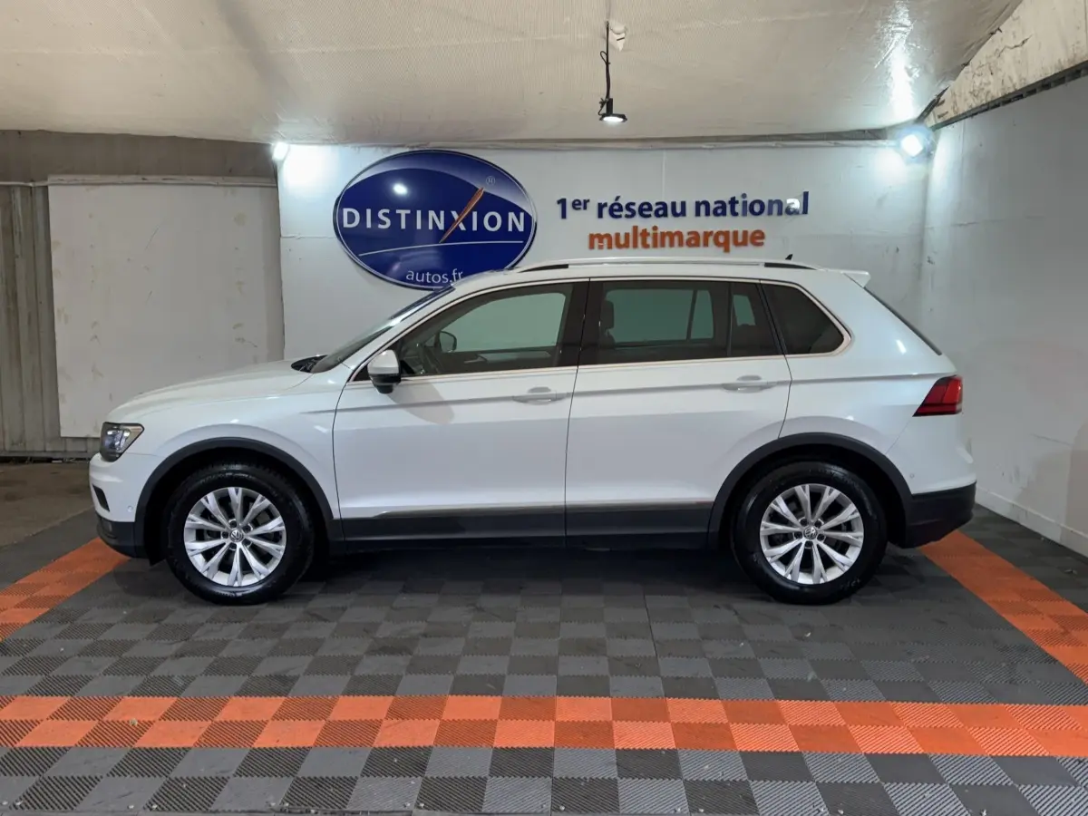 Profil côté gauche d'un Volkswagen Tiguan blanc 2020 stationné en intérieur avec jantes alliage et barres de toit noires.