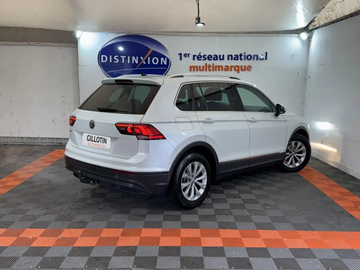 Vue 3/4 arrière droite d'un Volkswagen Tiguan blanc stationné en intérieur avec barres de toit noires et jantes argentées.