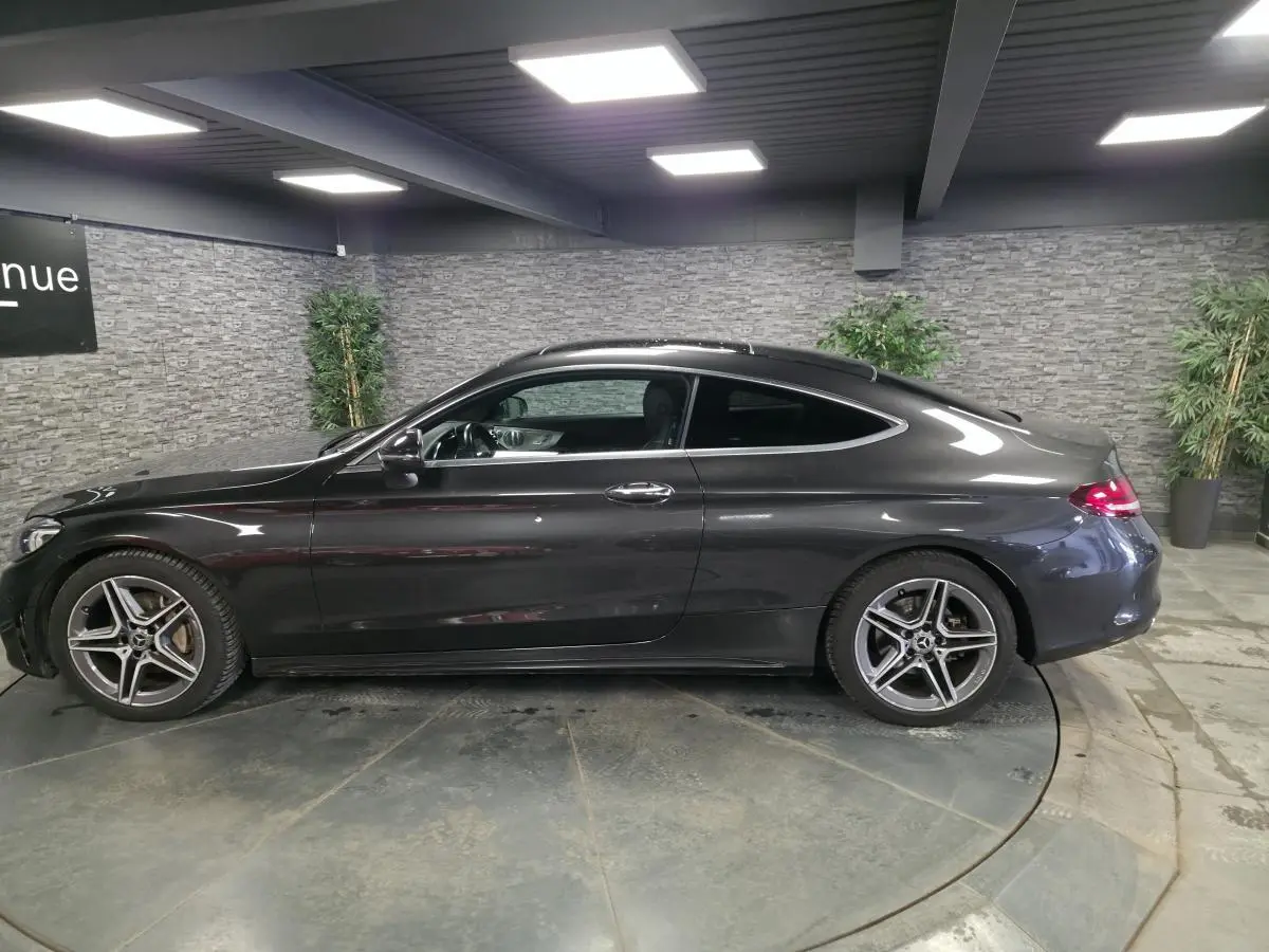 Vue de profil côté gauche d'un coupé Mercedes Classe C gris métallisé avec jantes AMG 18 pouces en intérieur showroom.