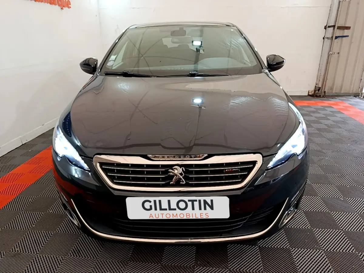 Vue avant d’une Peugeot 308 gris foncé avec phares allumés et calandre chromée dans un garage.