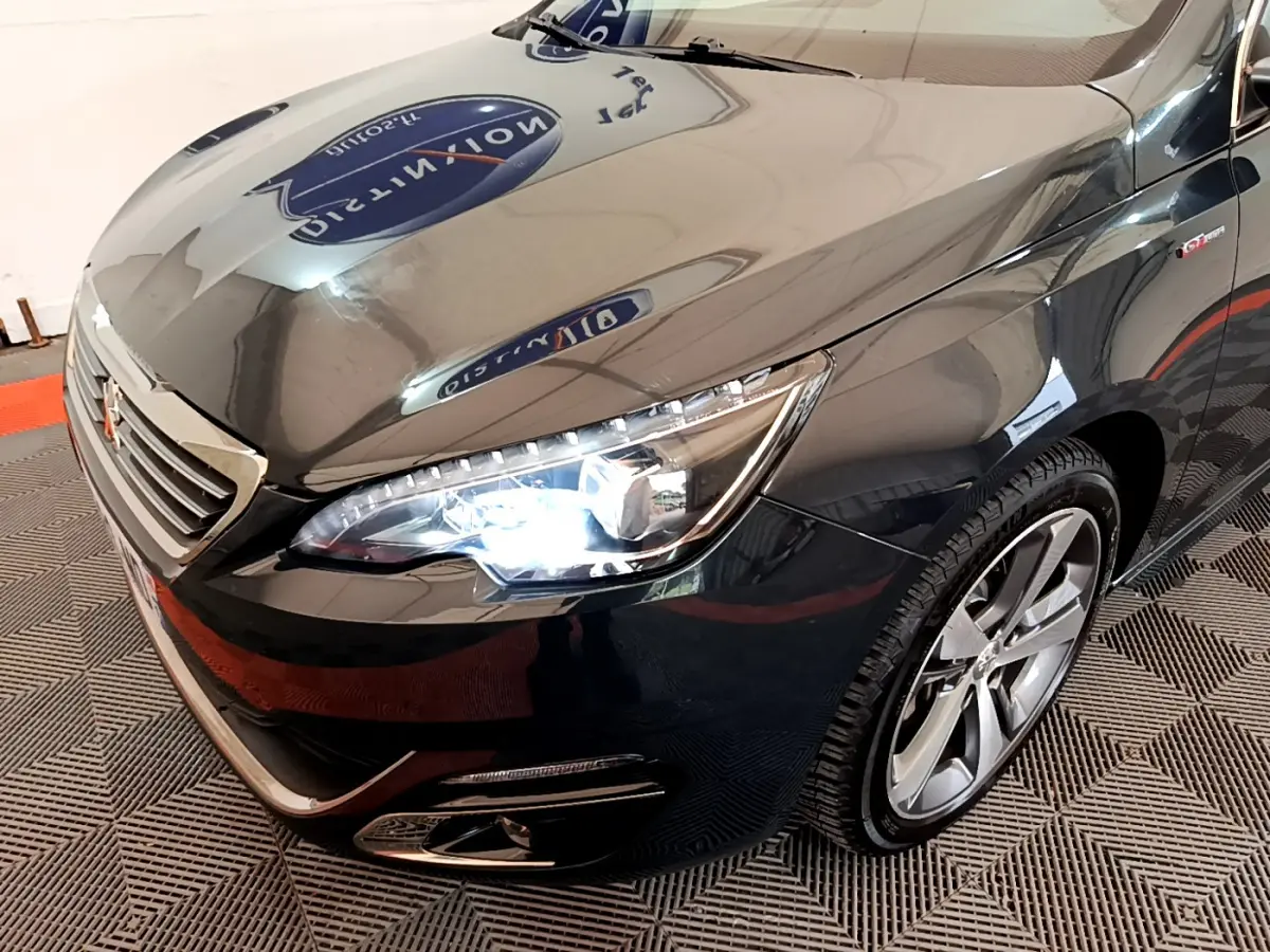 Vue 3/4 avant droit d'une Peugeot 308 gris foncé avec phares allumés et jantes alliage visibles.