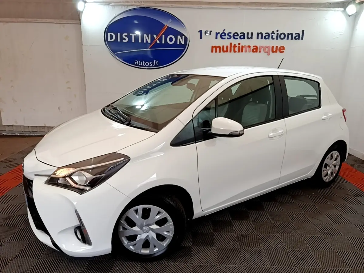 Toyota Yaris blanche vue 3/4 avant droit, avec phares allumés et jantes en alliage visibles.