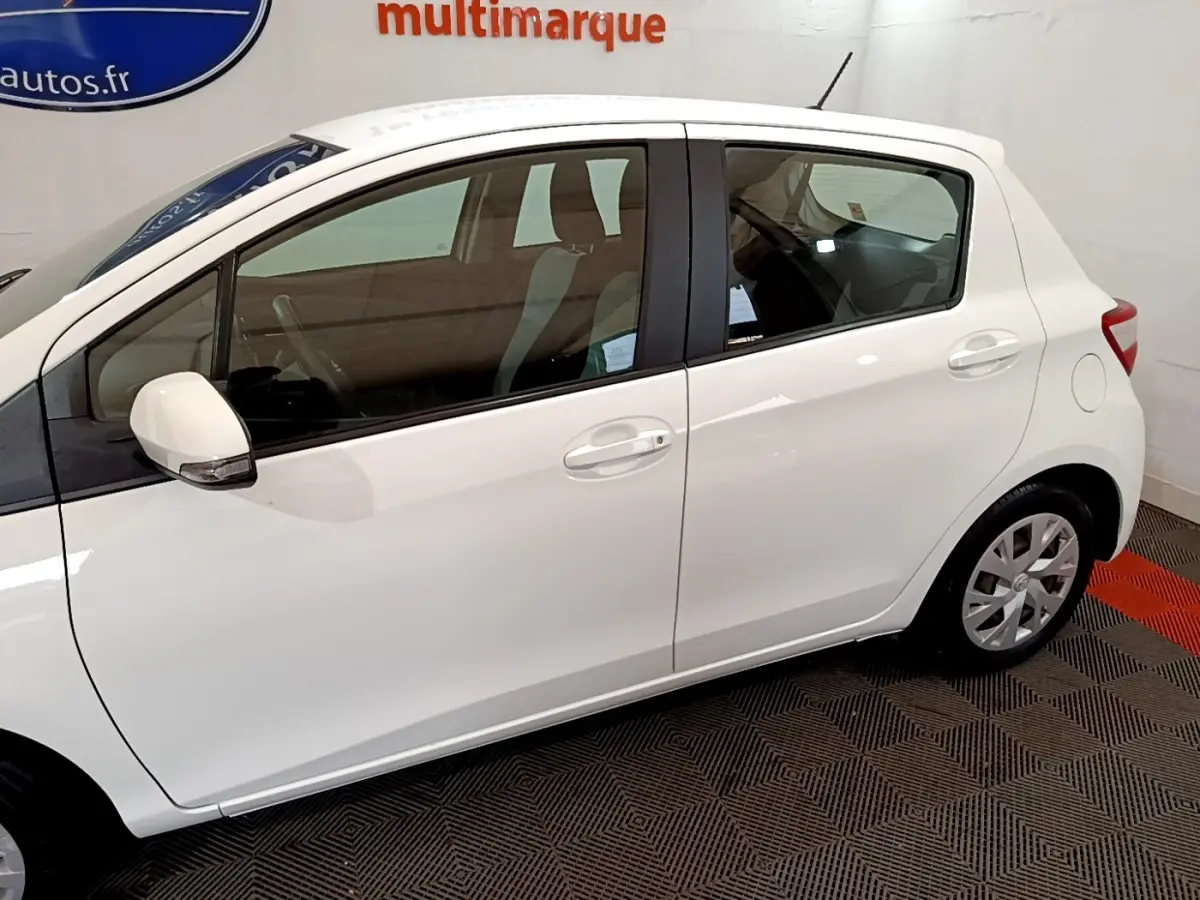 Profil droit d'une Toyota Yaris blanche 2019 en version hybride essence, visible en intérieur sur sol damier noir et rouge.