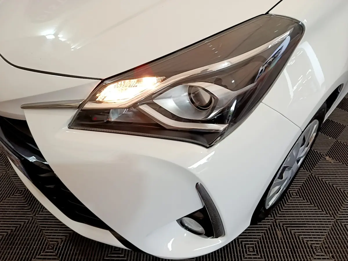 Gros plan sur le phare avant droit allumé d'une Toyota Yaris blanche, avec partie de la calandre et roue visible.