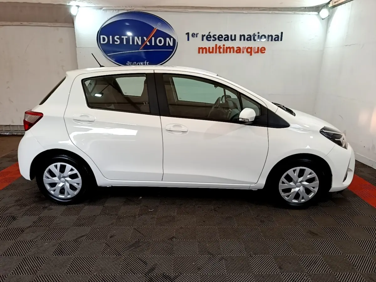 Profil droit d'une Toyota Yaris blanche 2019 essence hybride, vue en intérieur showroom avec sol à motifs.