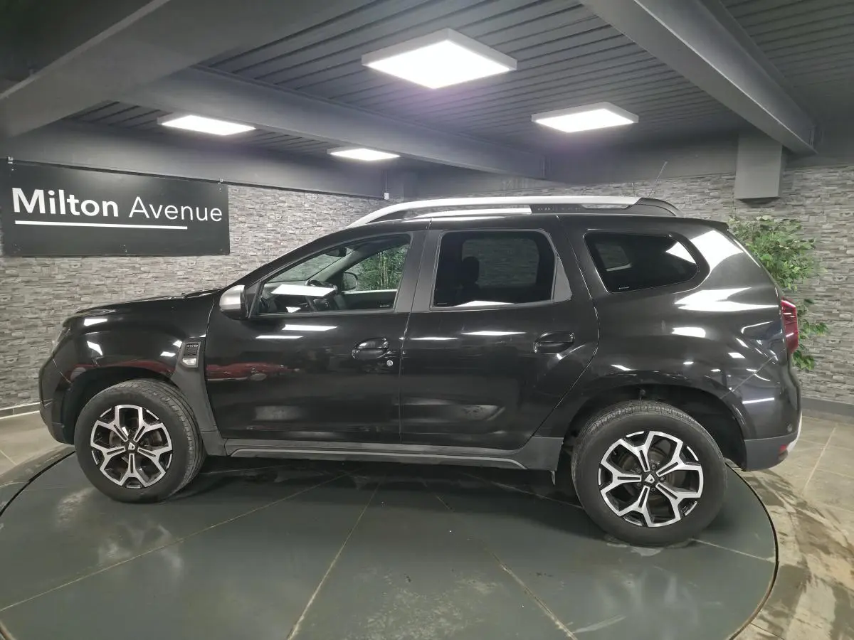 Profil droit du Dacia Duster noir nacré 2019 avec jantes alliage 17 pouces et barres de toit visibles en intérieur showroom.