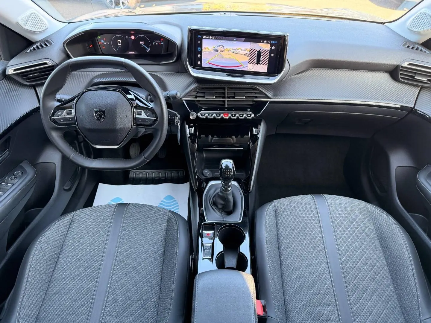 Vue intérieure avant de la Peugeot 208 2025, tableau de bord et volant compact avec écran tactile et levier de vitesse manuel.