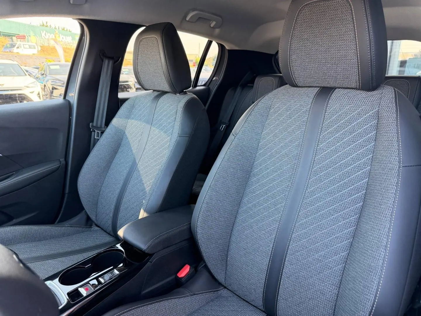 Vue intérieure montrant les sièges avant gris tissu et cuir de la Peugeot 208 1.2 100ch Allure 2025, avec accoudoir central.