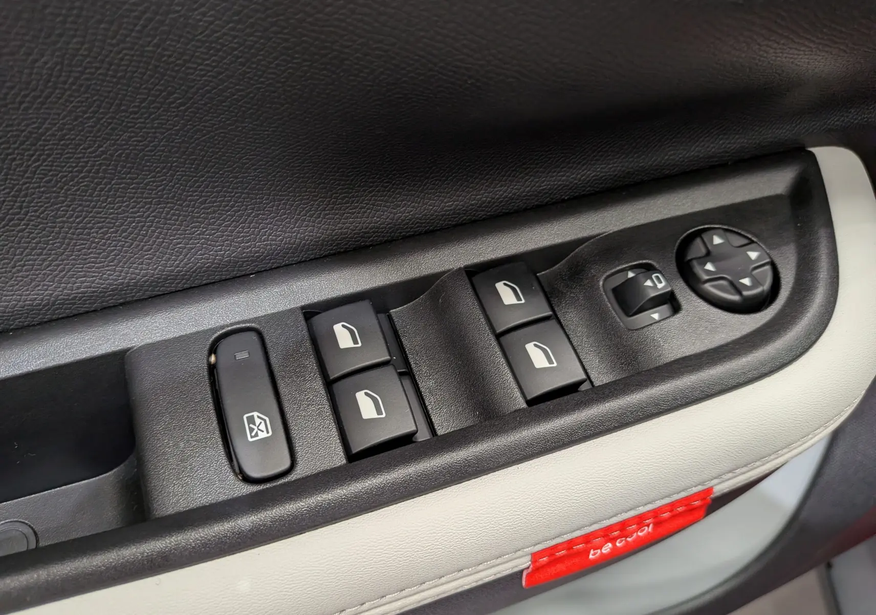 Gros plan sur les commandes des vitres et rétroviseurs côté gauche d’une Citroën C3 Aircross Hybride 2026 avec garniture beige et étiquette rouge.