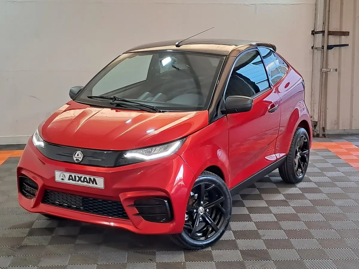 Vue 3/4 avant d'une Aixam Ambition rouge 2025 avec toit noir et jantes noires brillantes dans un intérieur neutre.