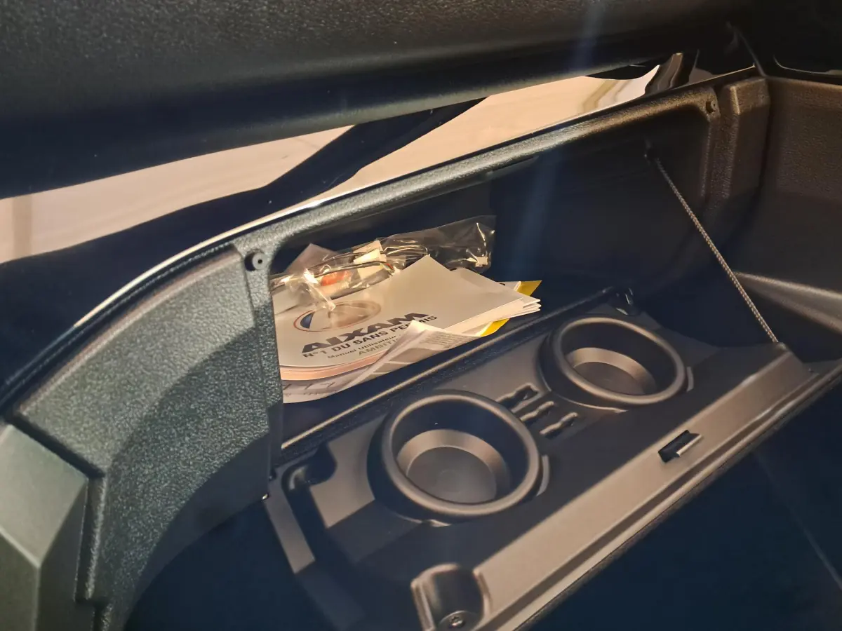 Gros plan sur la boîte à gants ouverte d'une Aixam Ambition rouge, avec porte-gobelets et documents visibles à l'intérieur.