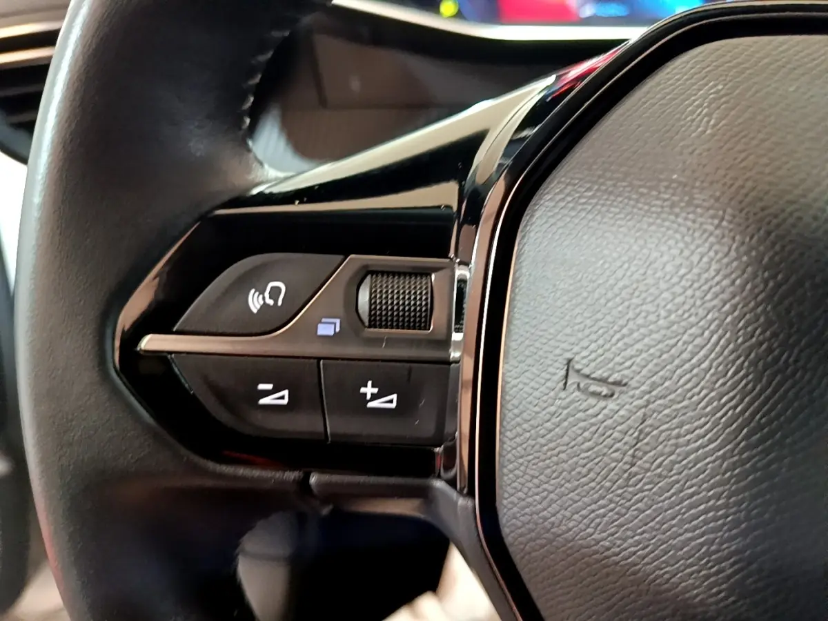 Gros plan sur les commandes gauche du volant cuir noir de la Peugeot 208 blanche, version PureTech 100 S&S Allure Business.