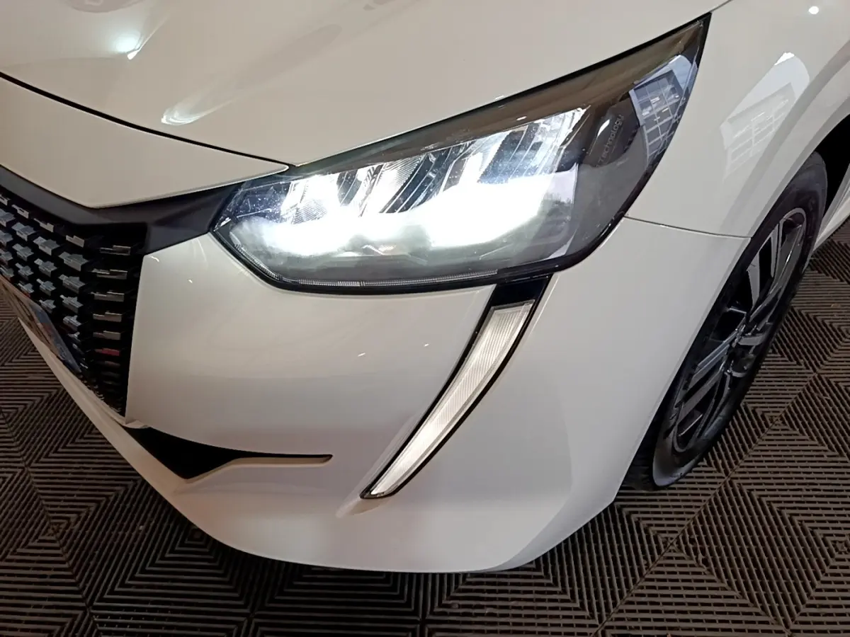 Gros plan sur l'avant droit blanc d'une Peugeot 208 2021, mettant en valeur le phare LED allumé et la calandre noire.